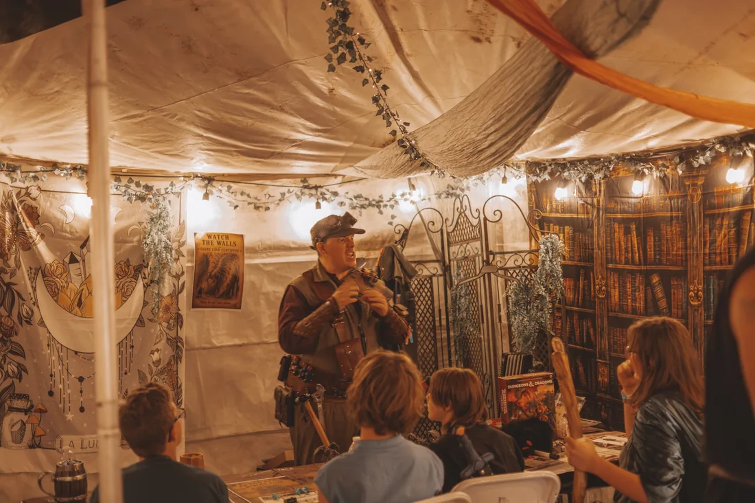 A costumed game facilitator leading a storytelling adventure for participants around a table in a warmly decorated tent.